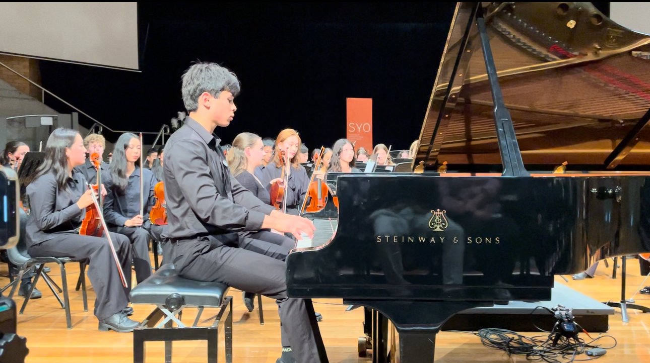 A young man playing a Steinway & Sons grand piano during an orchestra performance with an audience in the background.