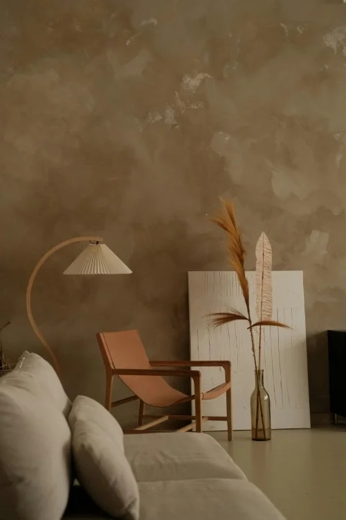  A  living room corner featuring a hand-troweled Textured Finish walls in a warm taupe, styled with a tan leather lounge chair, curved wooden floor lamp, and minimalist white canvas. 