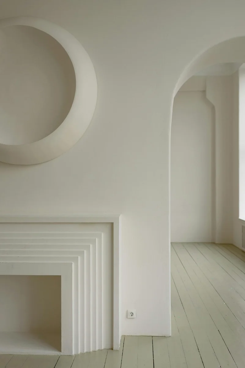A high-end minimalist interior featuring seamless white microcement wall finishes, a sculptural stepped fireplace, and an elegant arched doorway in an organic modern style.