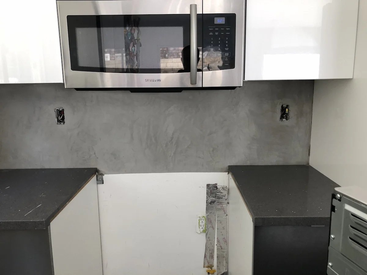 A kitchen backsplash area featuring a seamless concrete plaster finish installed between white upper cabinets and dark lower cabinets.