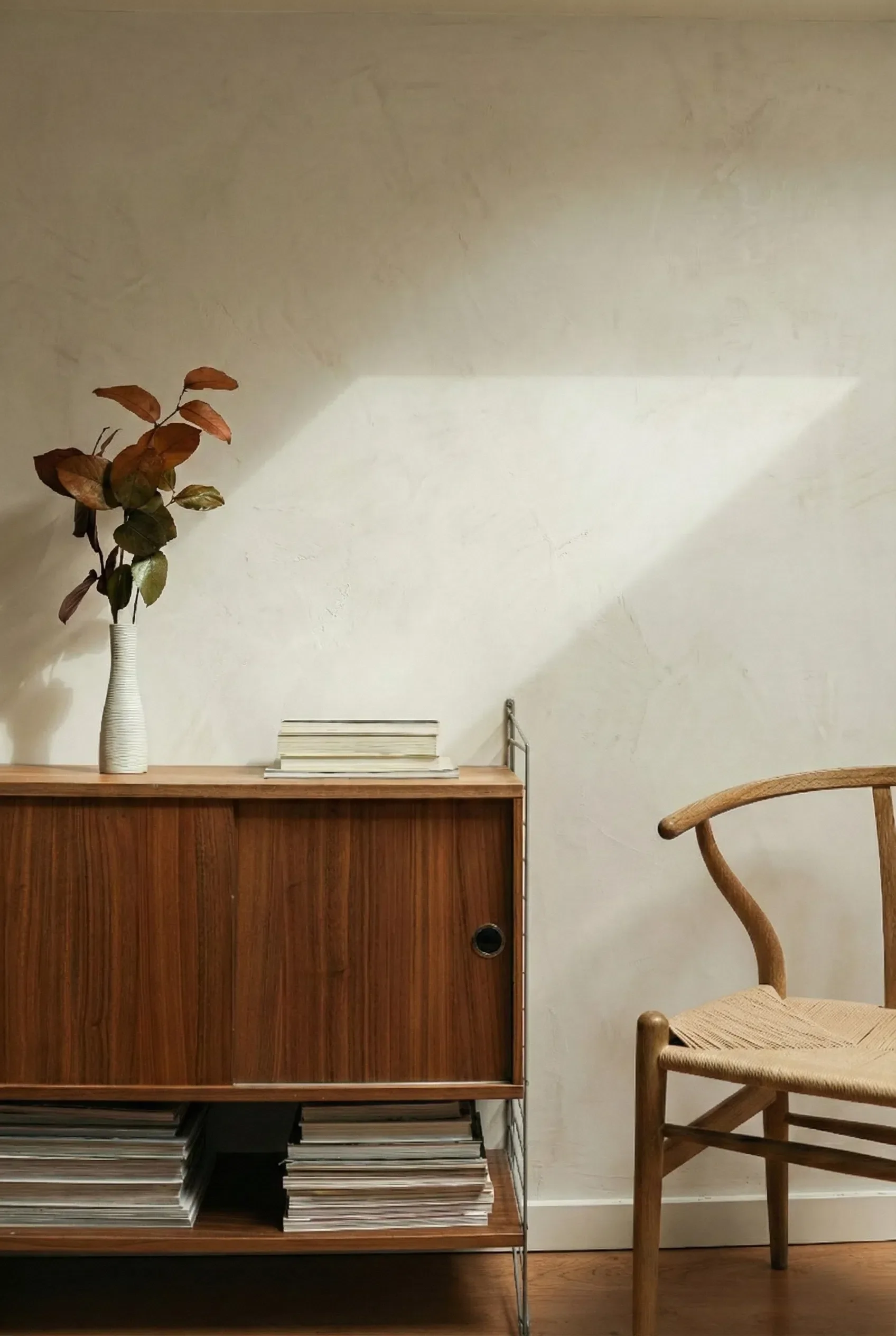 Sunlight streaming across a beige Venetian plaster wall, highlighting the artisanal trowel marks in a quiet, minimalist corner with mid-century modern wood decor.