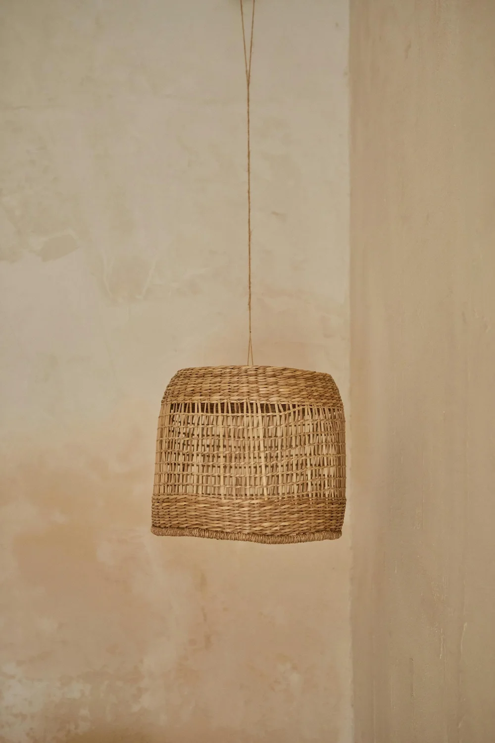 Detailed corner view of a matte Venetian plaster wall in warm beige tones, featuring subtle organic movement and a soft mineral texture, styled with a hanging woven rattan pendant light.
