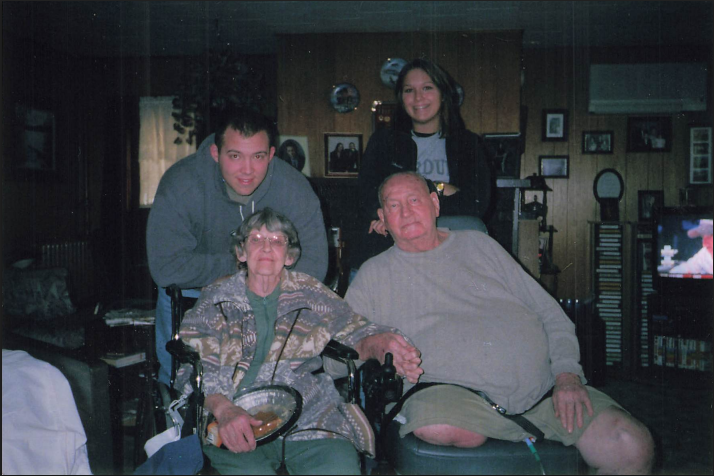 A family photo inside a living room featuring four people: an elderly woman and man seated, with the woman in a wheelchair, and a young man and woman standing behind them, all smiling.