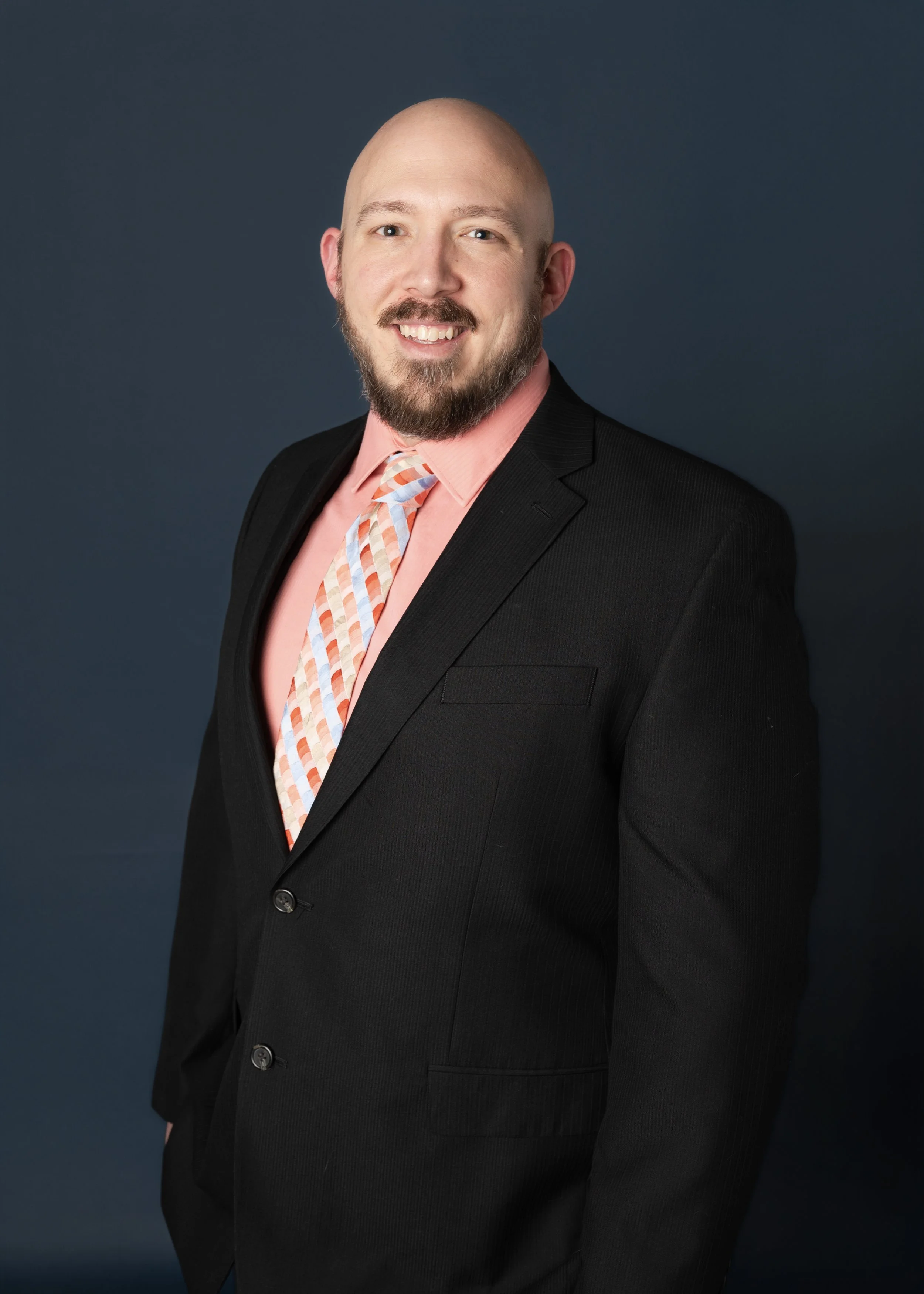 A man with a beard and mustache, smiling, wearing a black suit jacket, a pink shirt, and a colorful striped tie, standing against a dark blue background.