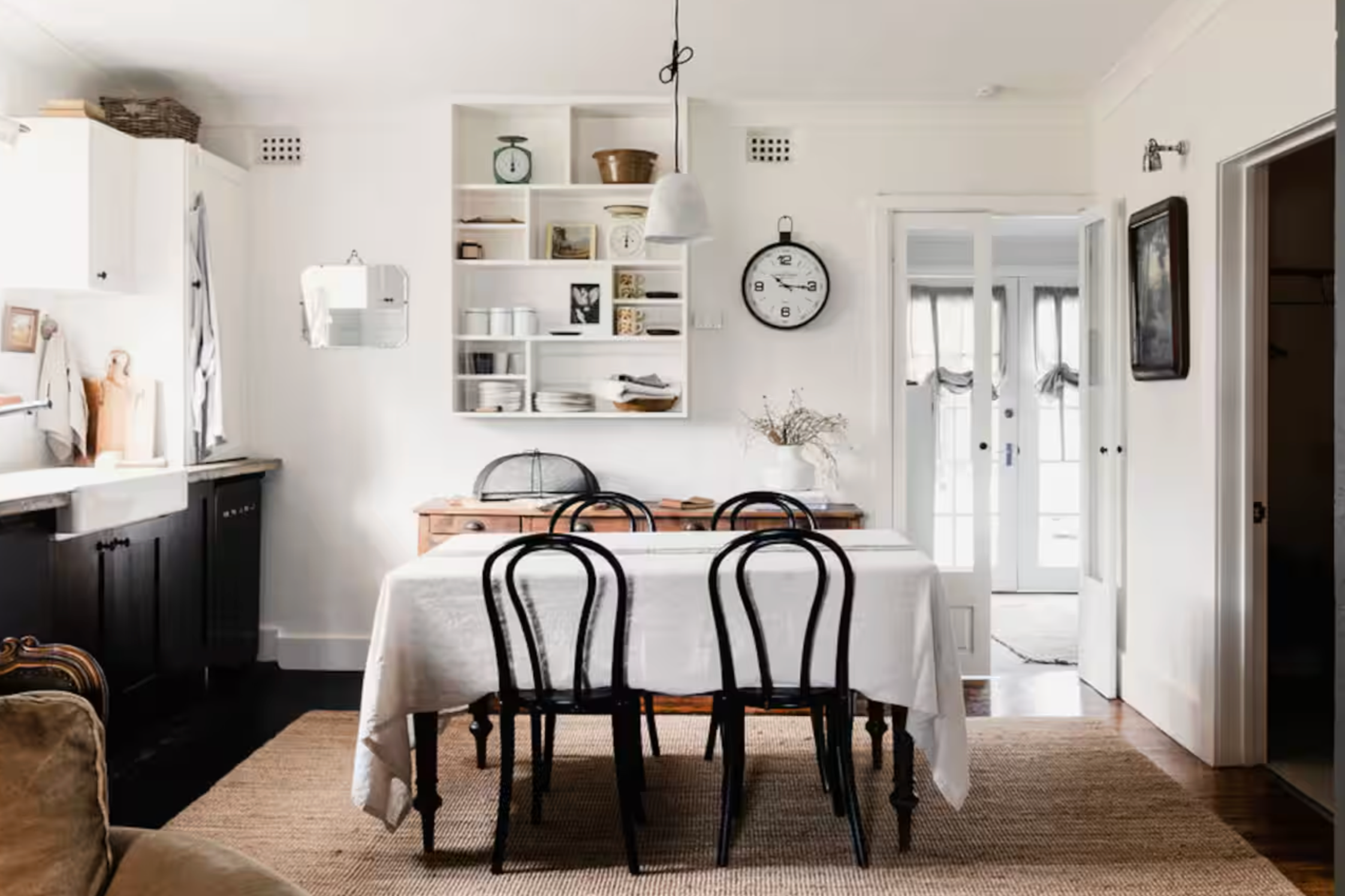 Dining room with a wooden table covered with a white tablecloth, surrounded by black chairs, with a vase of dried flowers, and white walls with open shelves, a wall clock, and a doorway leading outside.