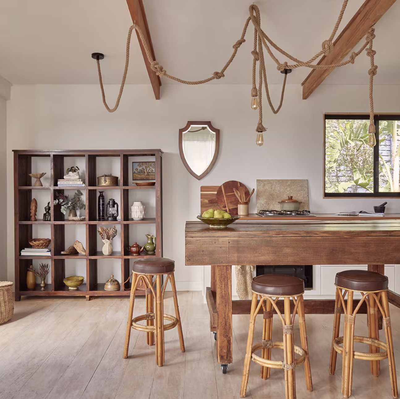 A cozy kitchen with a wooden dining table and four barstools, a wooden bookshelf filled with decorative items, and a rustic rope chandelier hanging from the ceiling.