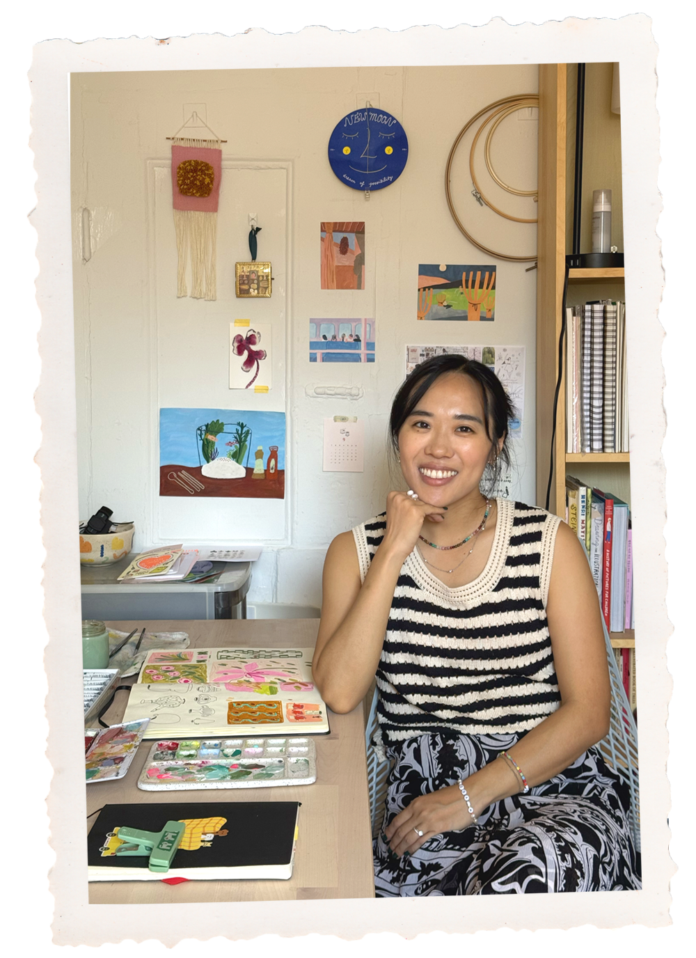 A woman sitting at a desk surrounded by art supplies with colorful artwork and decorations on the wall behind her.