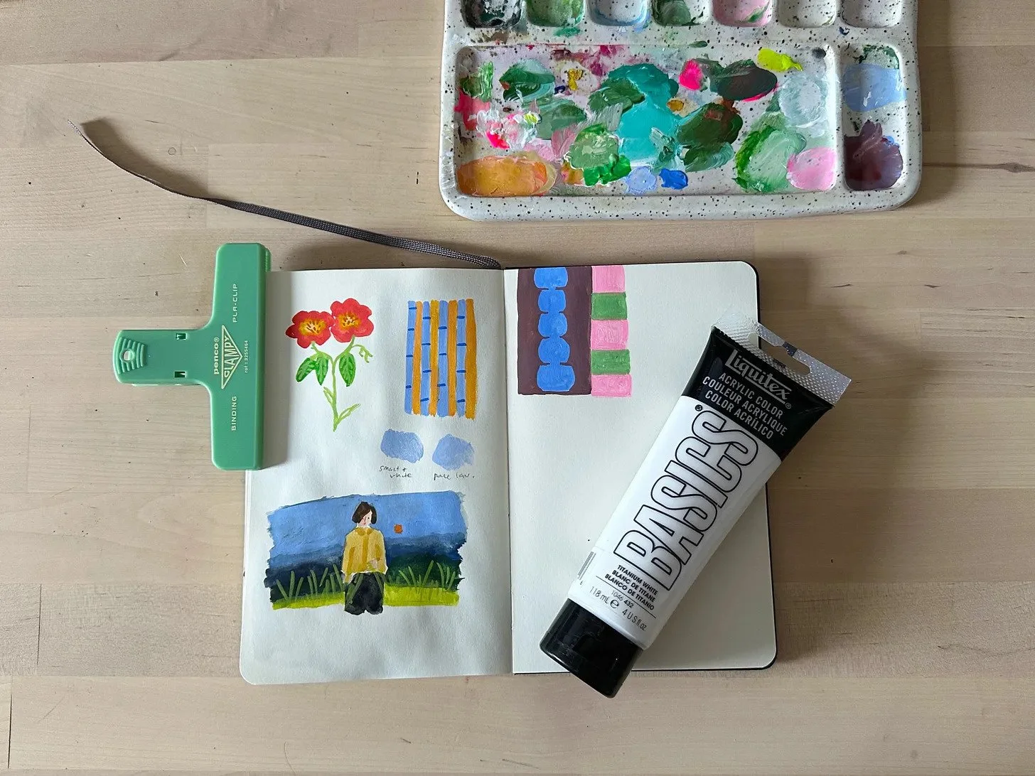 Open sketchbook with painted scenes, a tube of paint, and a watercolor palette arranged on a desk.