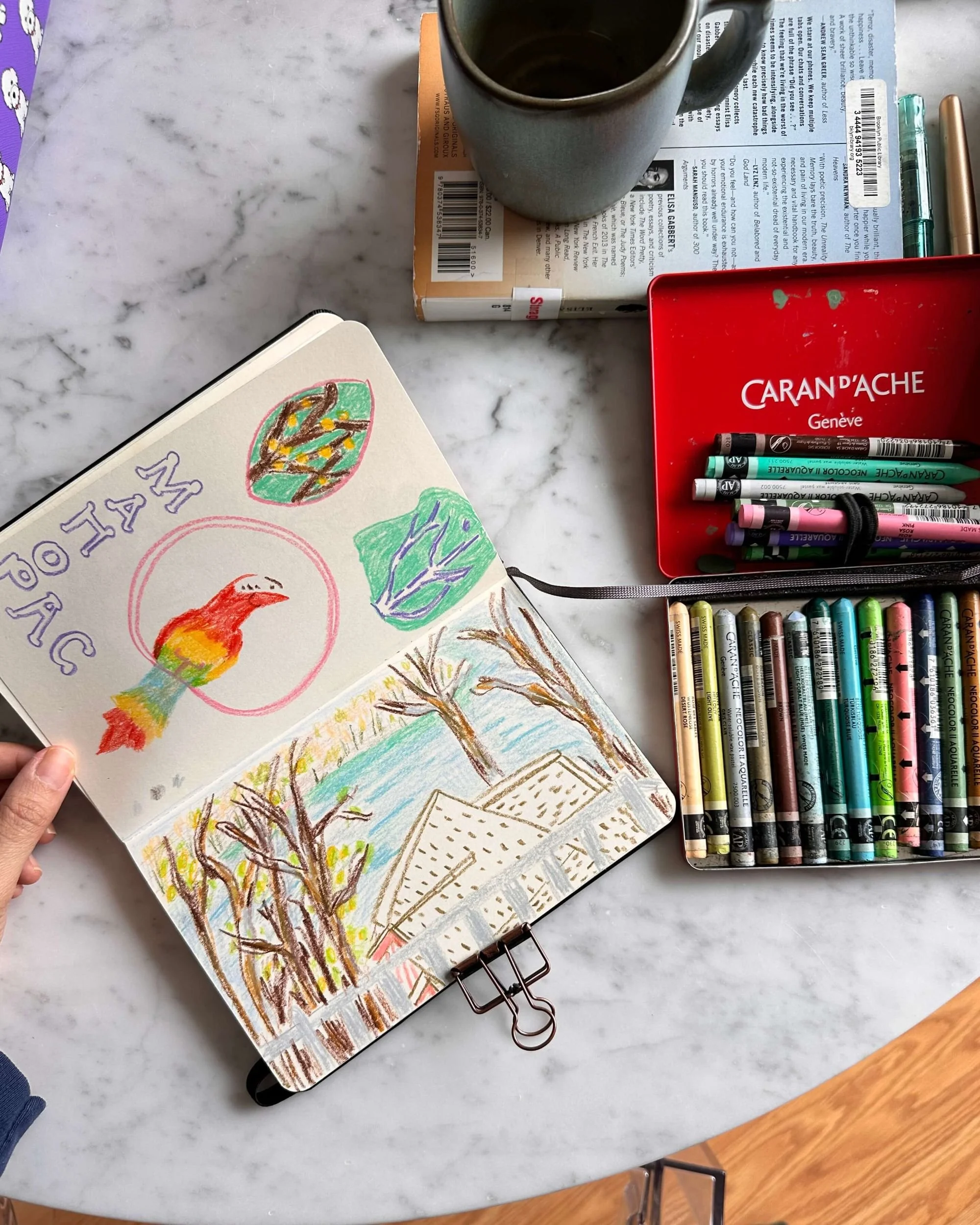 Open sketchbook with colorful drawings of a bird, trees, and a landscape on a marble table. Art supplies including markers, pens, and a red box are nearby. A coffee mug and a book are also on the table.
