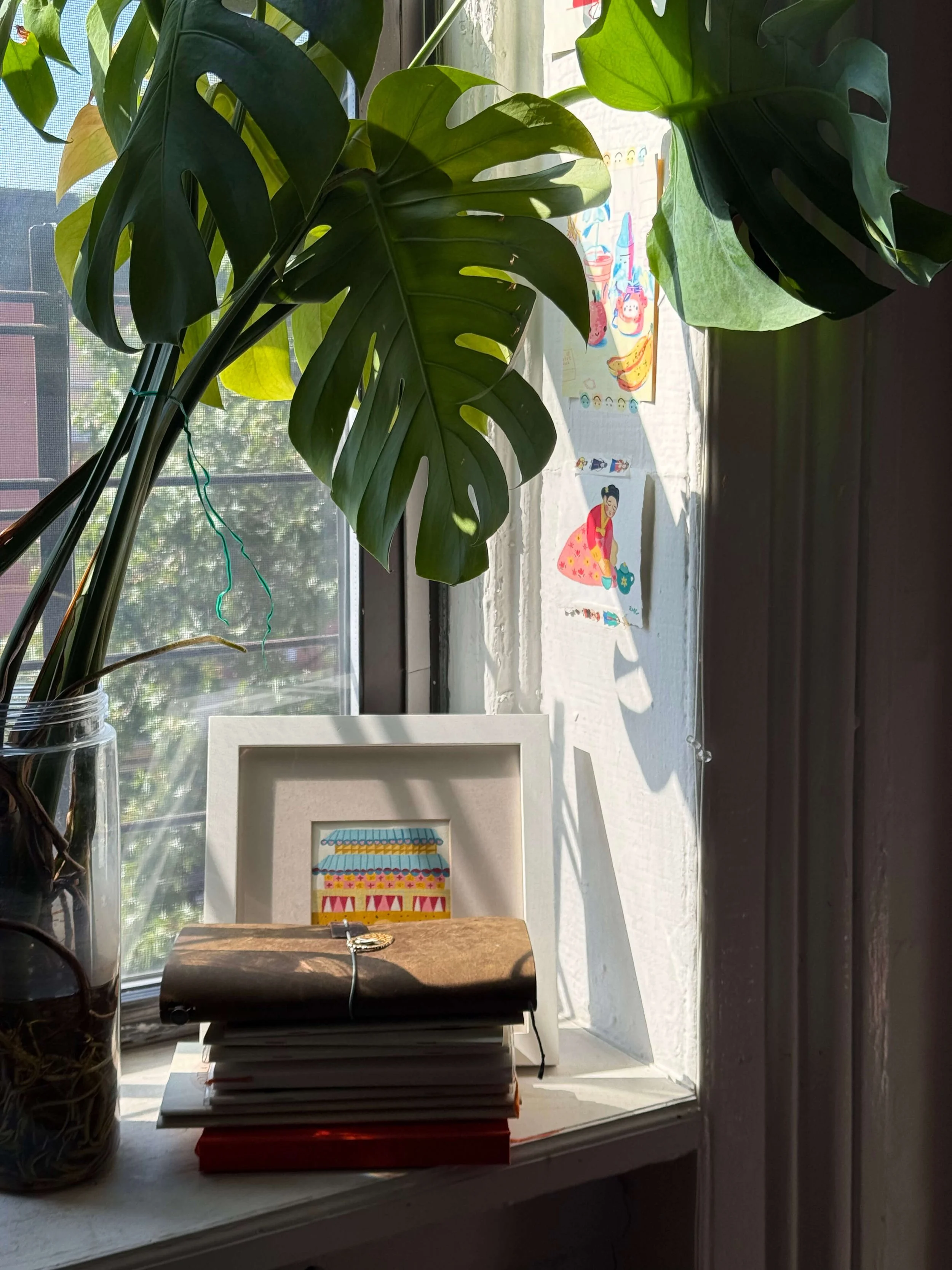 Stack of Carolyn Yoo’s sketchbooks on a windowsill beside framed art and houseplants.