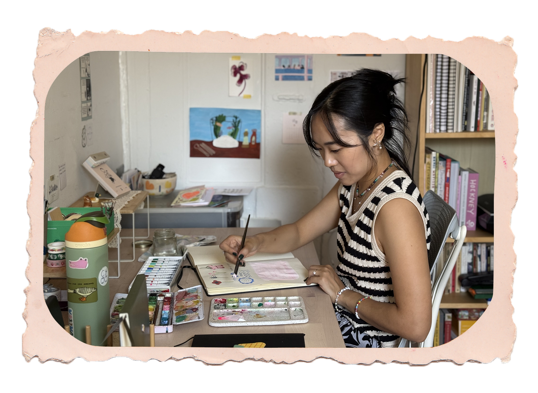 Carolyn Yoo painting in her sketchbook at her desk, surrounded by art materials