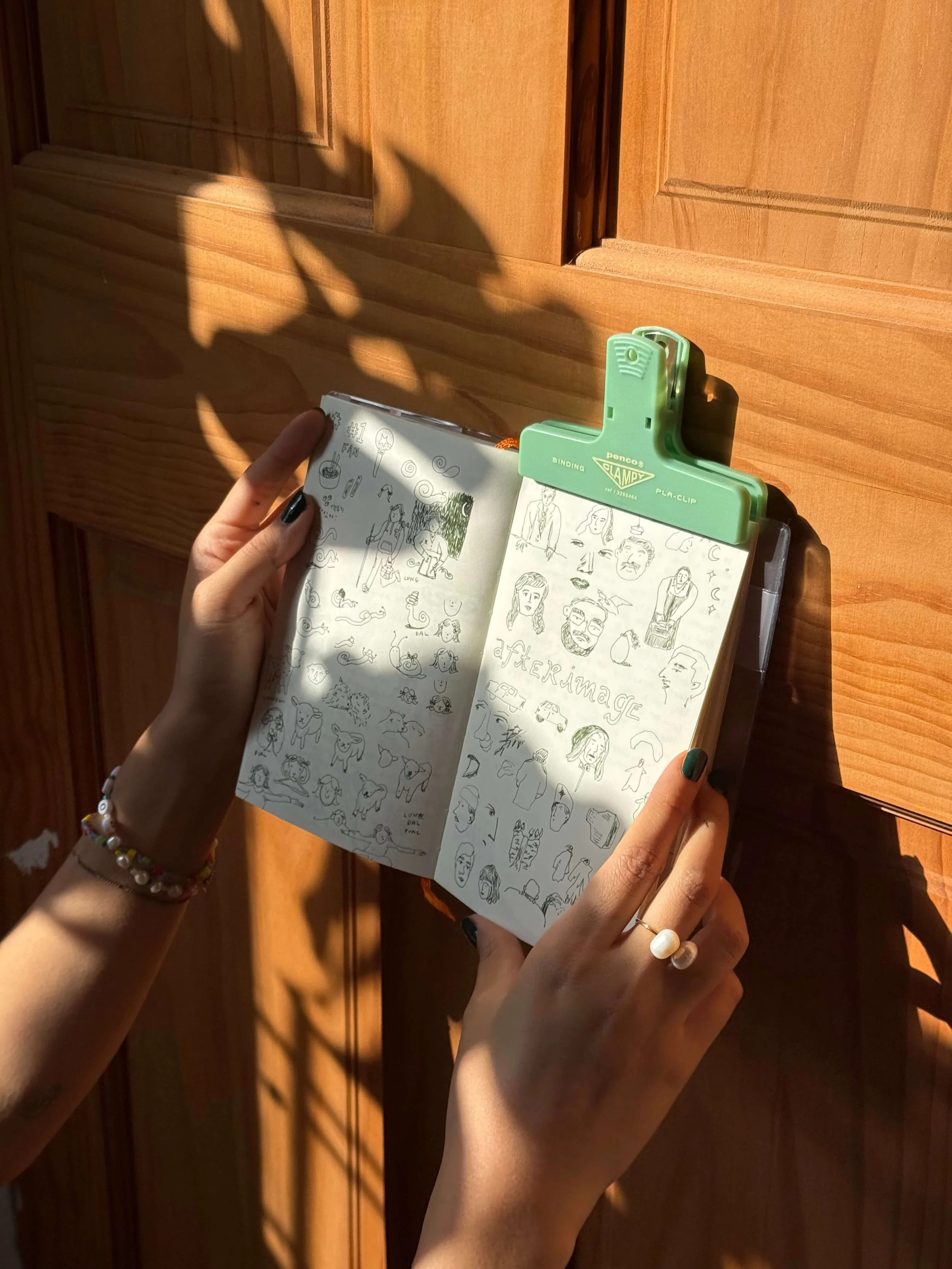 Close-up of a person's hands holding an open sketchbook with black ink sketches of faces, animals, and doodles, clipped to a wooden wall with a green binder clip, as sunlight creates shadows on the wall.