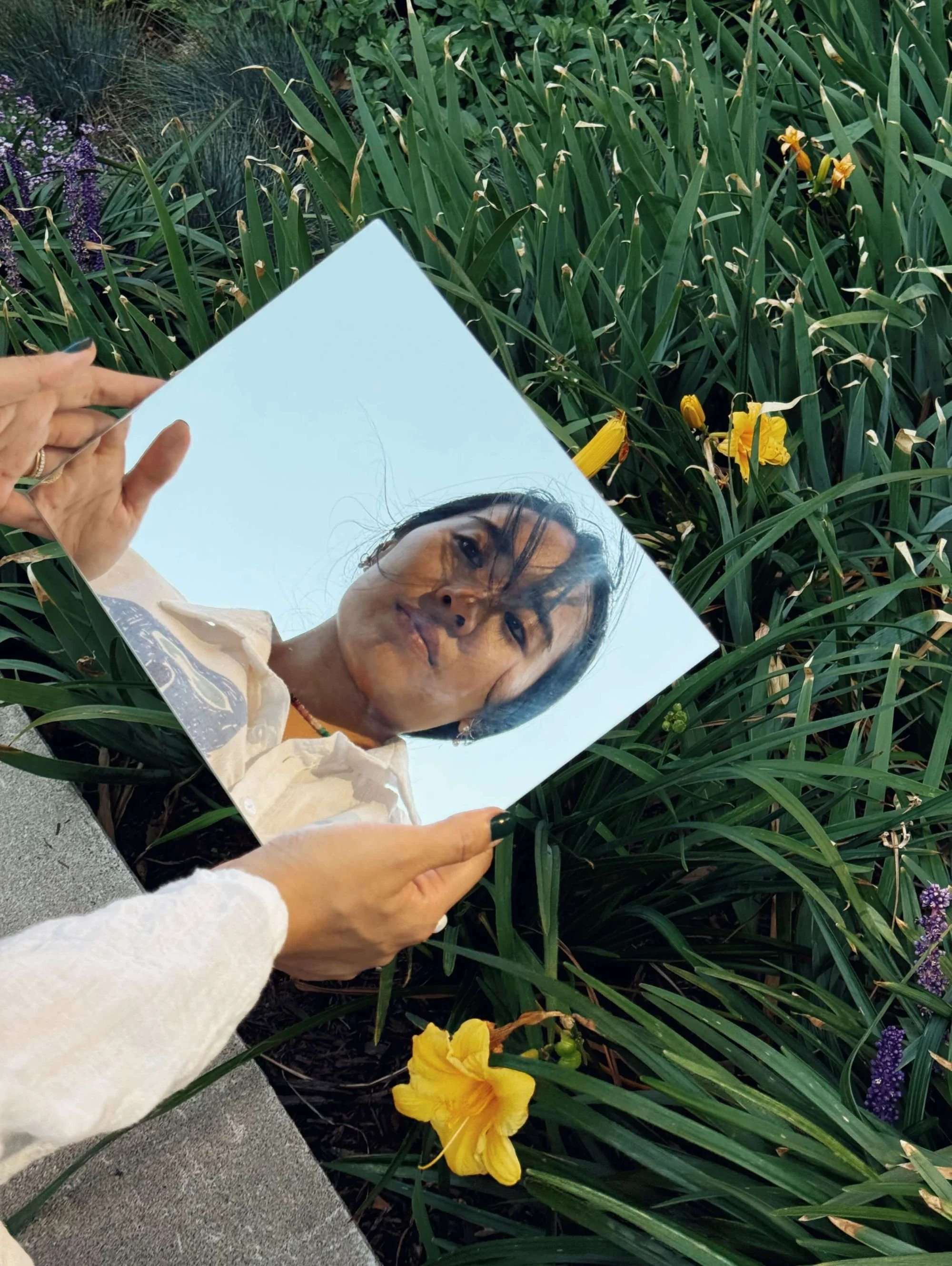 Carolyn Yoo holding a mirror outdoors, reflecting her face among flowers and greenery.