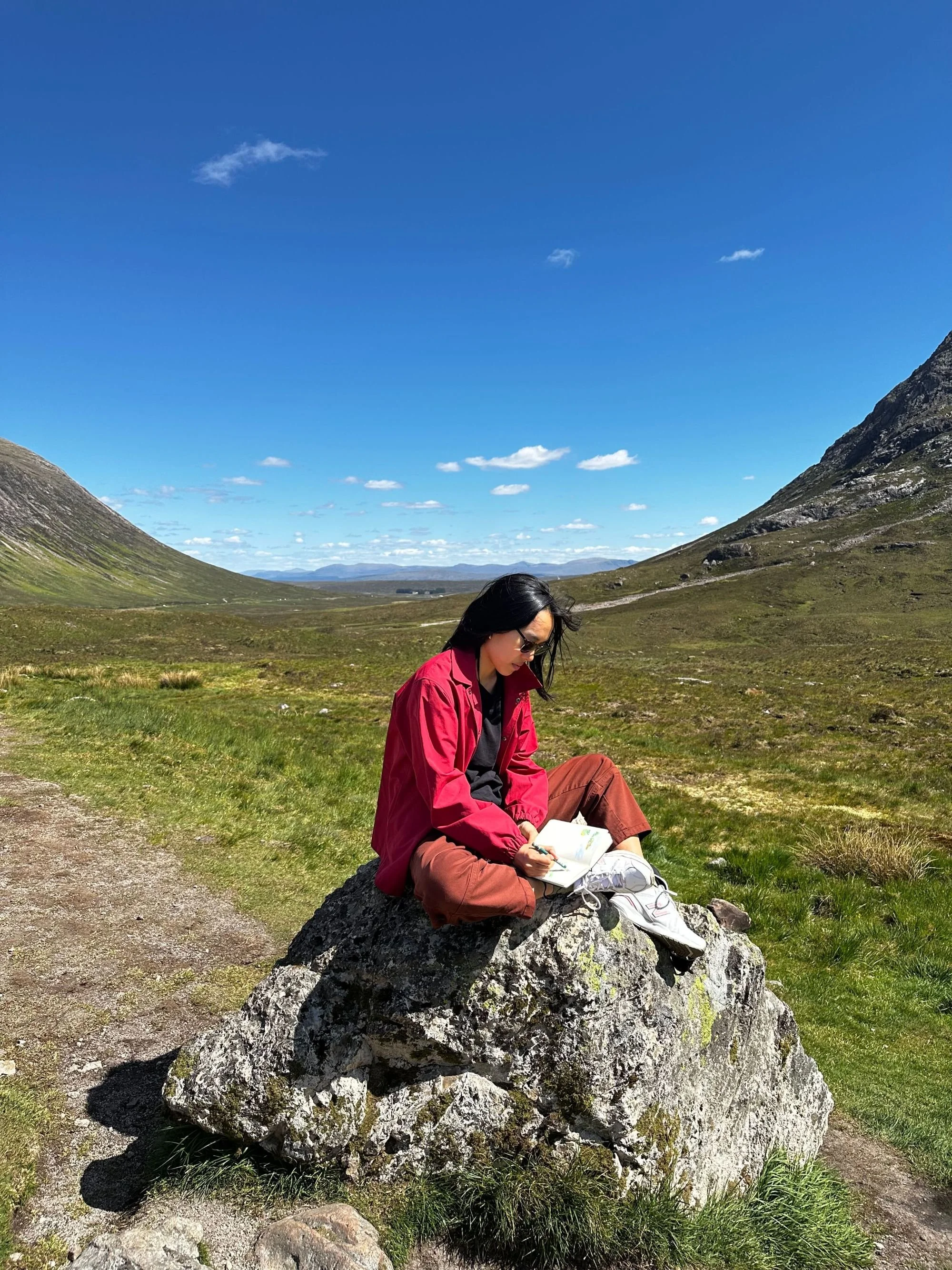 Carolyn Yoo sitting on a large rock outdoors, sketching in her notebook during a hike.