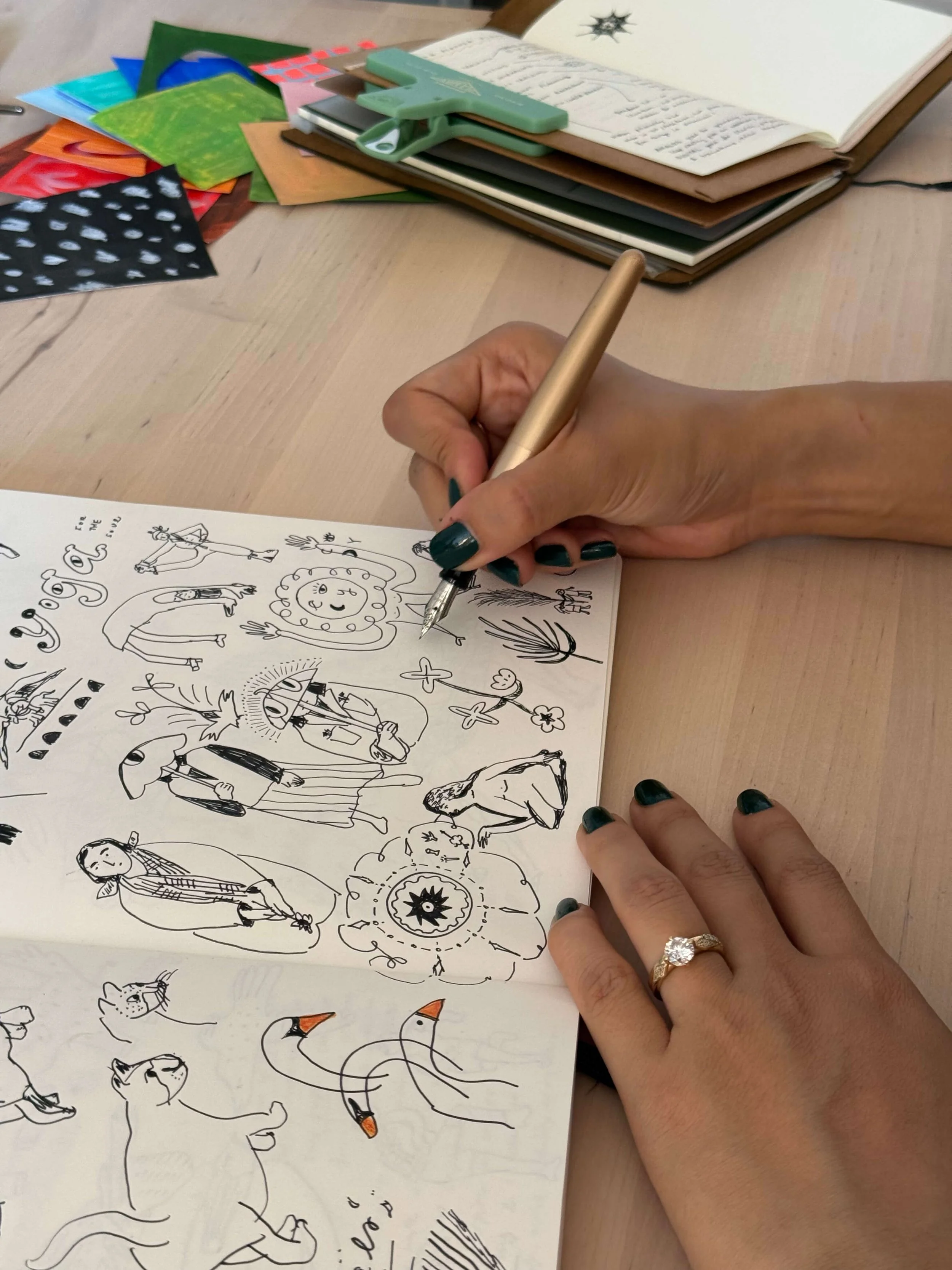 Person drawing black ink sketches of animals, people, and abstract designs on a white paper with a gold pen. On the table, there are colorful papers, a green binder clip, and a closed notebook.