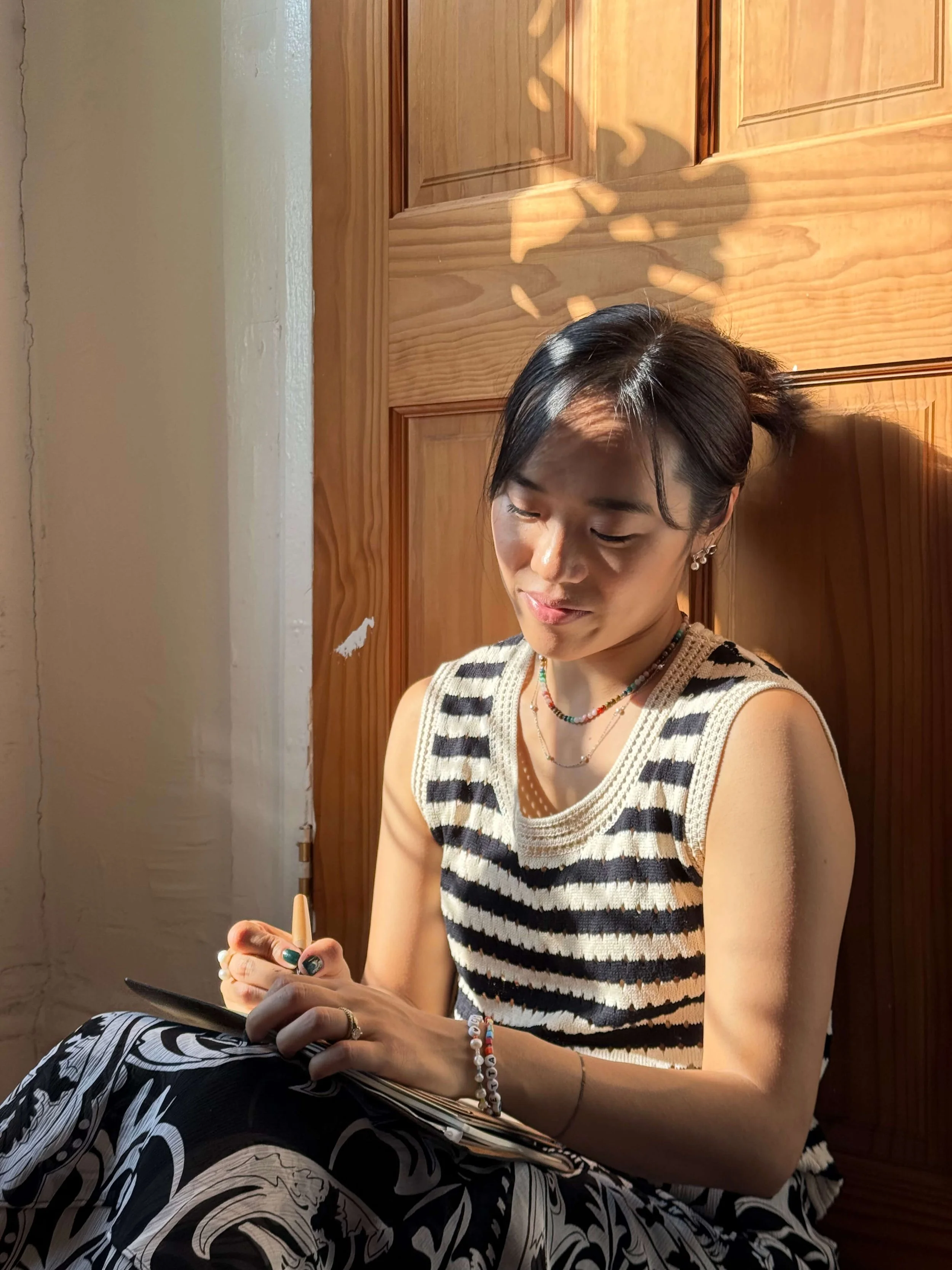 Carolyn Yoo sitting in sunlight by a wooden door, writing in her notebook.