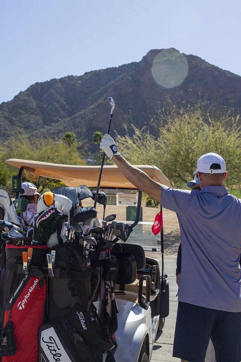 A man in a blue shirt and white cap standing next to a golf cart filled with golf clubs, raising a golf club in front of a mountainous desert landscape with sparse trees.