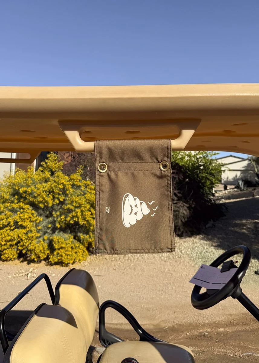 A brown fabric under the seat of a golf cart, with a white cartoonish face and steaming coffee cup printed on it, set outdoors with shrubs and trees in the background and a clear blue sky above.
