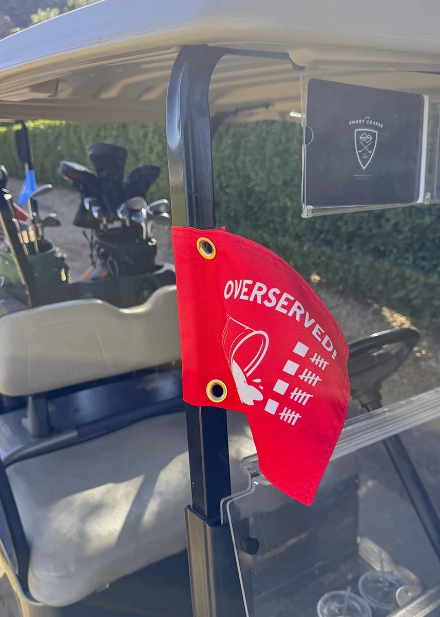 Red golf flag with the word "OVERSERVED" and check marks, attached to a golf cart, with golf clubs visible in the background.