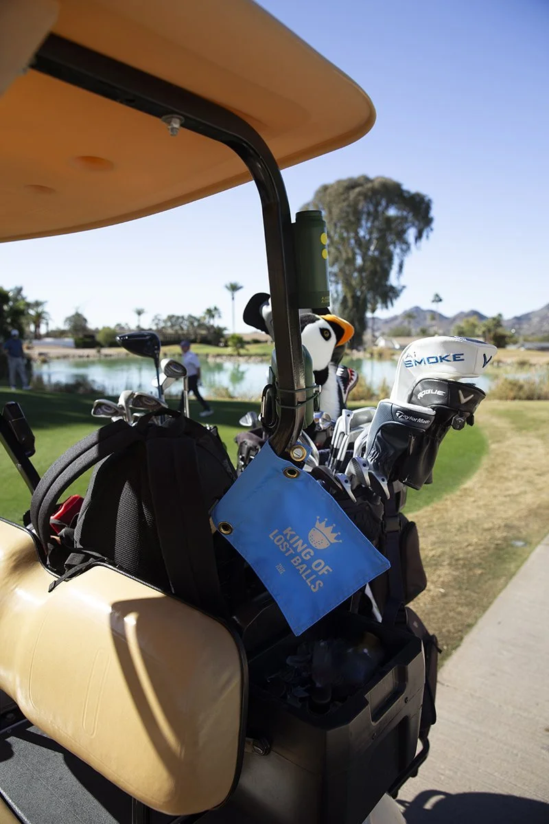 A golf cart parked on a golf course with golf clubs and a stuffed penguin toy in the back, near a pond and trees under a clear blue sky.