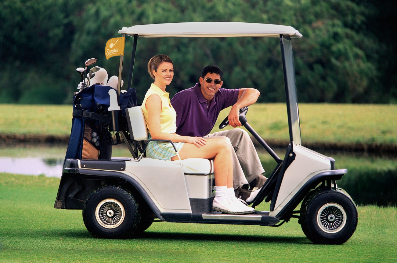 Two people are sitting in a golf cart on a golf course, smiling at the camera, with golf clubs and a Tally Flag in the back of the cart.