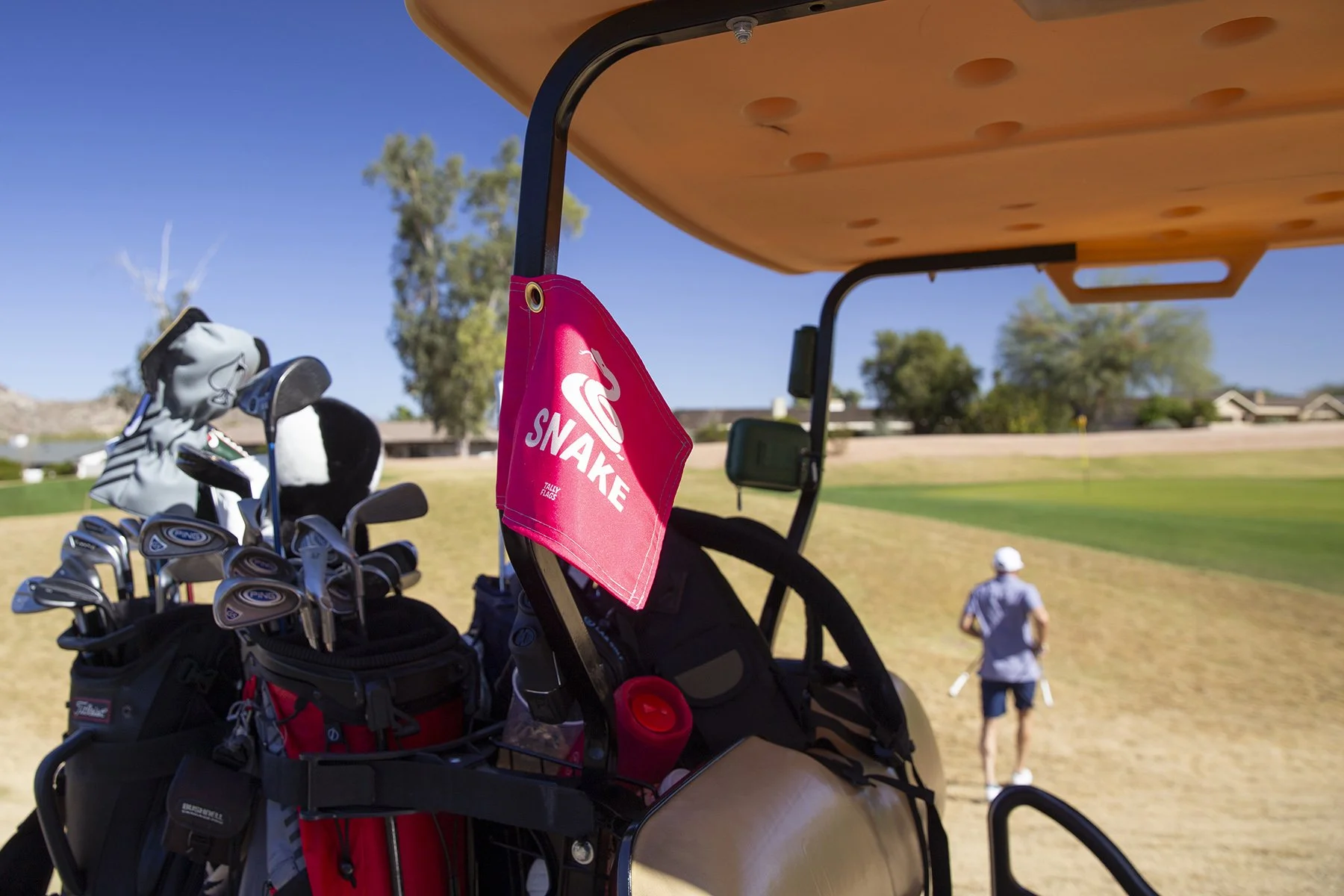 Golf cart with golf clubs and a pink towel hanging, view of a golfer walking on the golf course in the background with a clear blue sky.