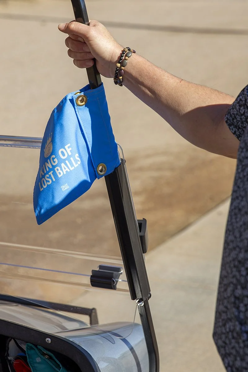 Person holding the handle of a luggage cart at an airport, with a blue bag attached that has white text reading 'KING OF LOST BALLS'.