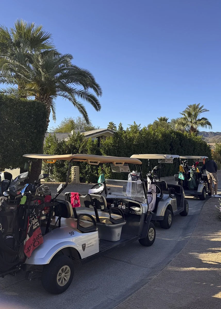 Several golf carts parked on a paved path, with golf bags and clubs loaded in the back, surrounded by palm trees and a bright blue sky.