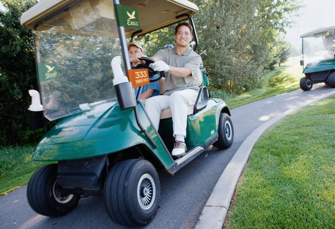 Two men riding in a green golf cart on a golf course pathway, with trees and grass surrounding them.