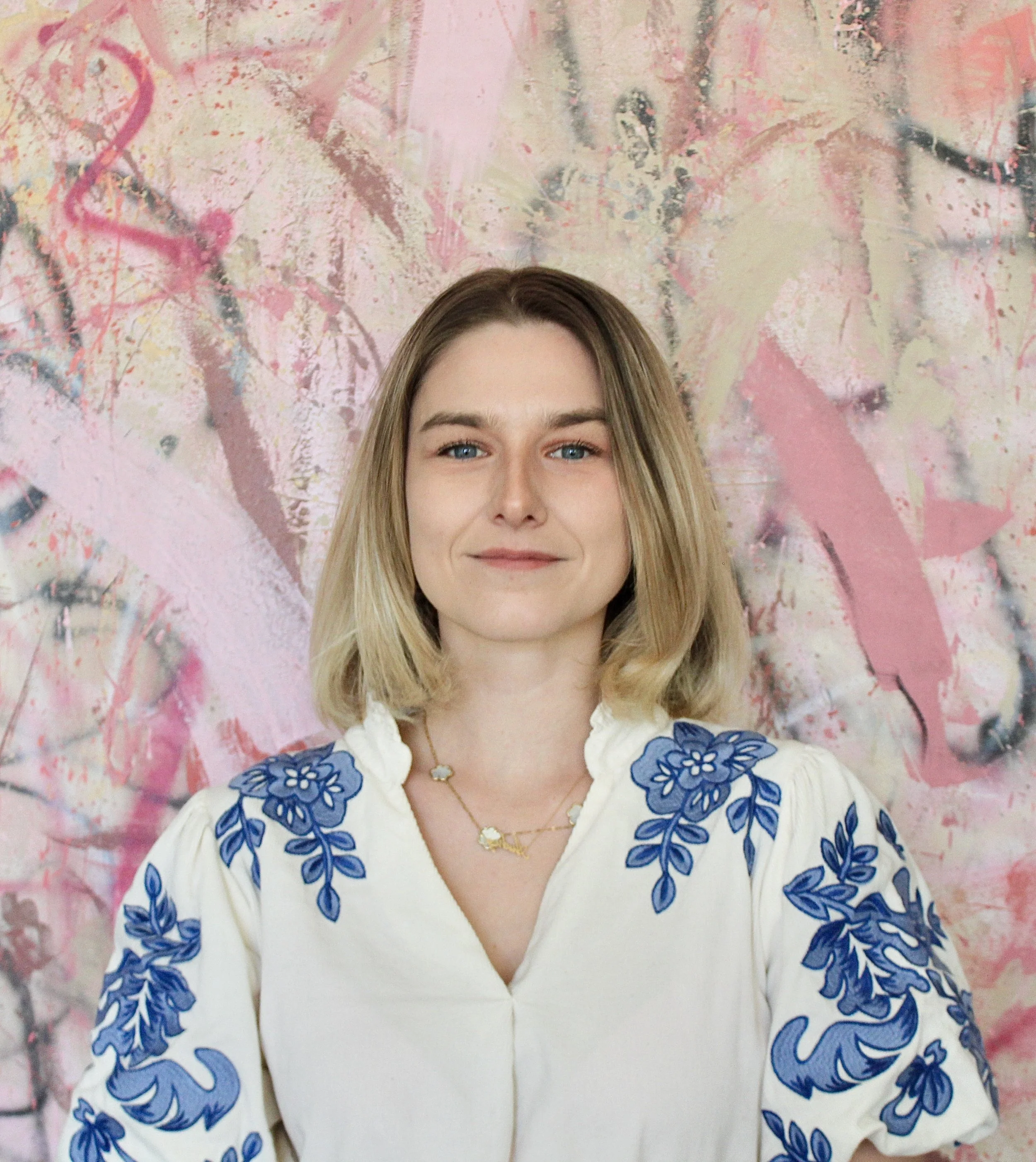 A woman with shoulder-length blonde hair, blue eyes, and fair skin, smiling slightly, dressed in a white blouse with blue floral embroidery on the shoulders and sleeves, standing in front of an abstract pink and pastel background.