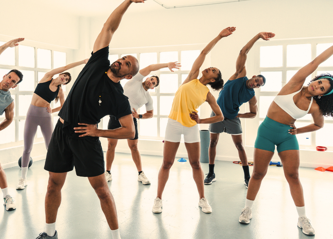 Group of diverse adults doing a stretching exercise in a fitness class, standing with one arm extended overhead and the other hand on their side.