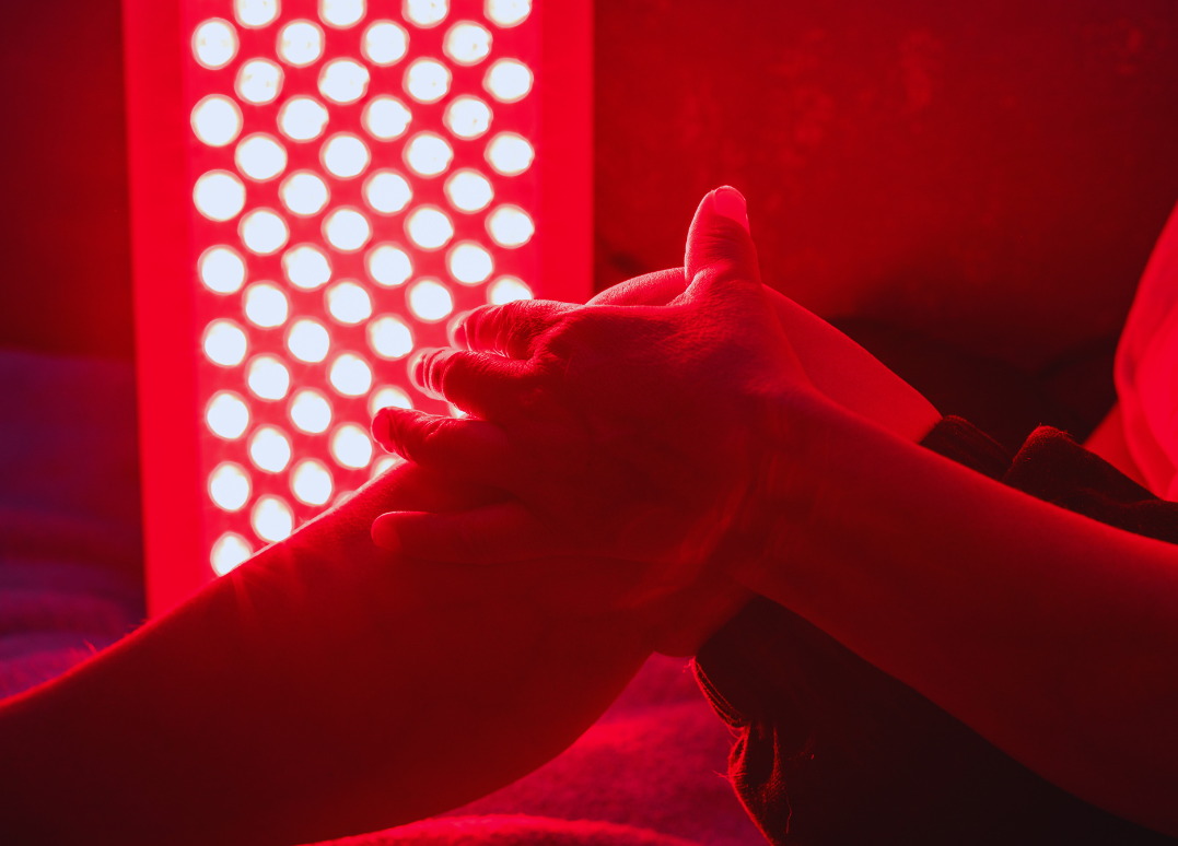 Two hands clasped together on a bed, illuminated by red light from a nearby LED therapy device.