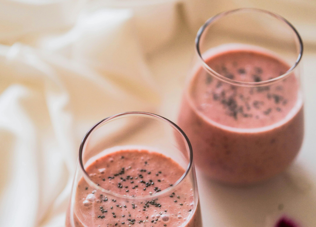Two glasses of pink smoothie topped with black chia seeds on a cream-colored surface with a soft-focus background.
