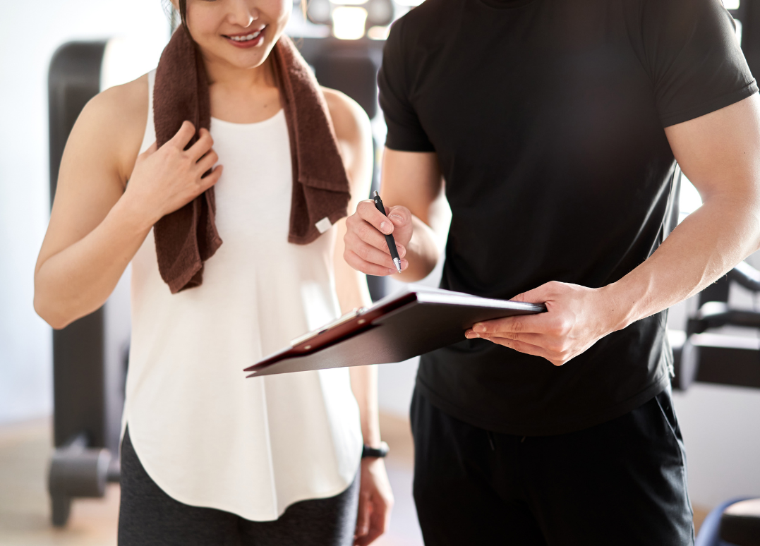 A woman and a man at the gym; the woman has a towel around her neck and is smiling, while the man is writing on a clipboard.