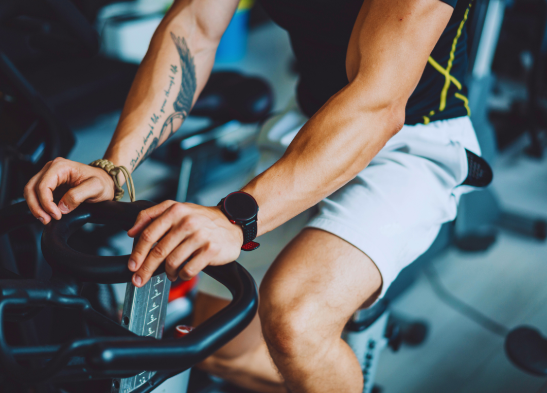 A person riding a stationary exercise bike in a gym, wearing a black smartwatch, a black sports shirt with yellow details, and white shorts.