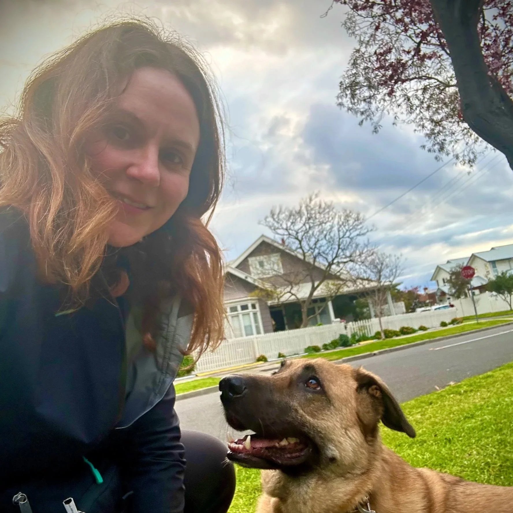 A woman with wavy brown hair smiling next to a brown dog with a happy expression on a grassy lawn in a suburban neighborhood with houses, trees, and a street in the background.