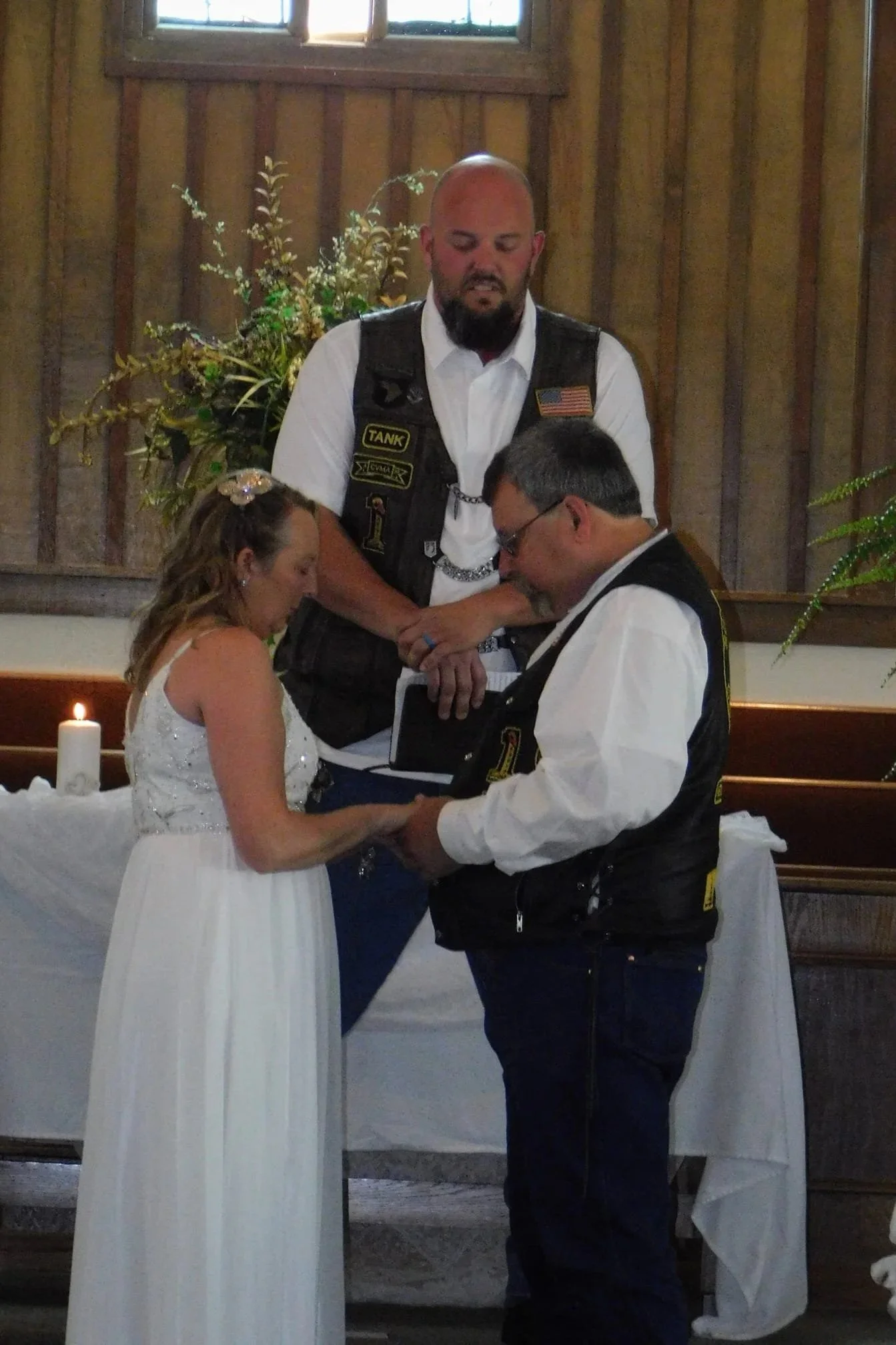 A wedding ceremony in a rustic wooden church with three people: a bride, a groom, and an officiant. The couple is holding hands and looking at each other, with the officiant standing behind them, observing. A lit candle and floral arrangement are in 