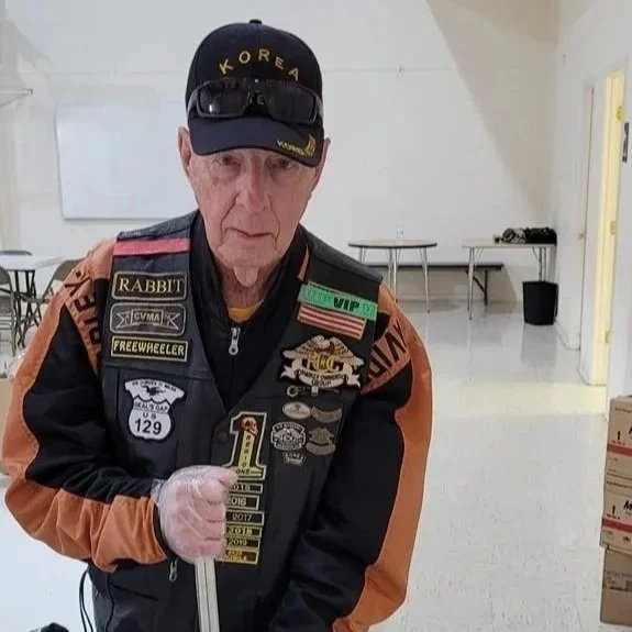 An elderly man wearing a black cap with 'KOREA' written on it, a black and orange jacket decorated with various patches and badges, and clear gloves, mixed chocolate in a large stainless steel pot using a long metal stirrer inside a bright, clean roo