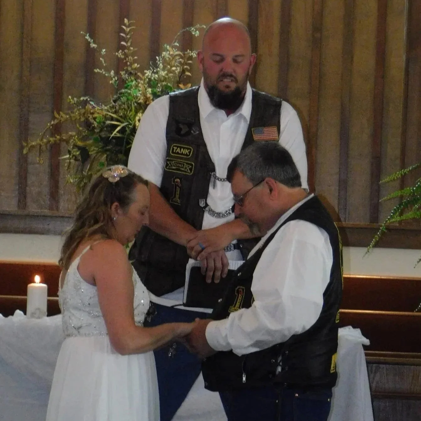 A wedding ceremony in a rustic wooden church with three people: a bride, a groom, and an officiant. The couple is holding hands and looking at each other, with the officiant standing behind them, observing. A lit candle and floral arrangement are in 