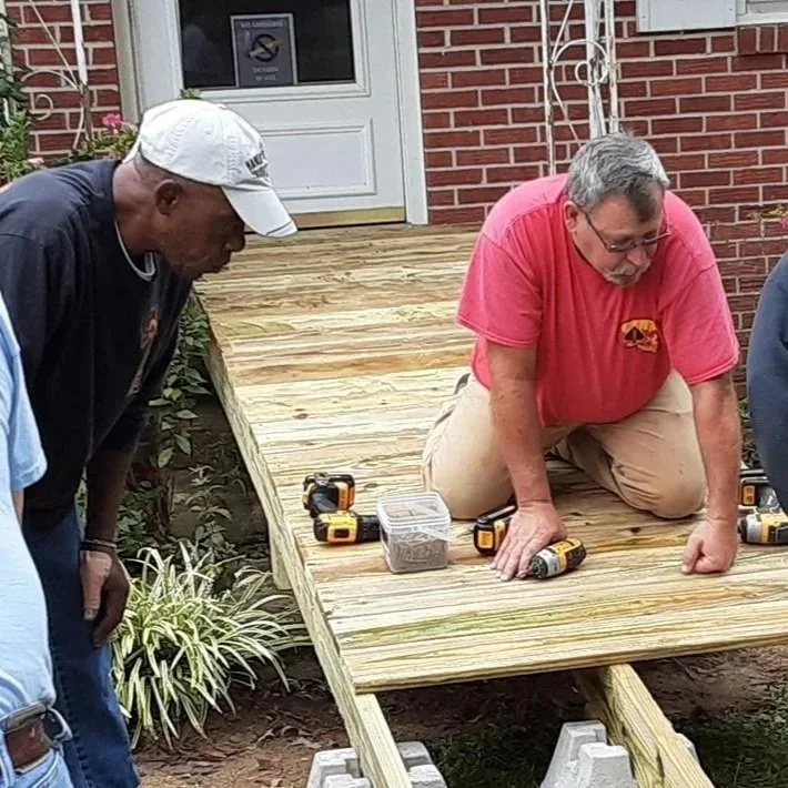 Four men working together to build a wooden ramp outside a brick house. Tools and materials are visible on the ramp, and the men are focused on the construction.