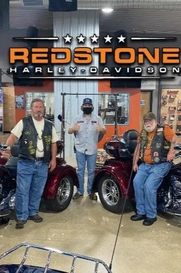 Three men standing inside a Harley Davidson dealership in front of a sign that says 'Redstone Harley-Davidson' with five stars above it. Two men are standing next to parked motorcycles, and one man is standing between them, giving a thumbs up. The men are wearing casual clothes and Harley Davidson vests, and one is wearing a mask.