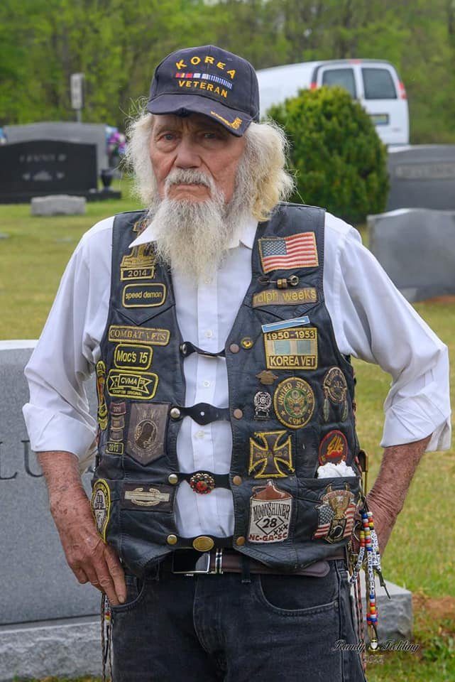 An elderly man with long white hair and a beard, wearing a black cap with 'KOREA VETERAN' embroidered on it, a white shirt, and a black vest covered with various military patches and badges, standing outdoors in a cemetery.