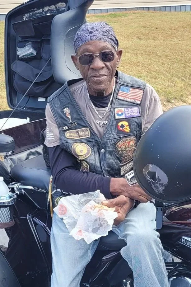 An elderly man wearing sunglasses, a bandana, and a leather vest with patches sitting on a motorcycle, holding a sandwich wrapped in paper.