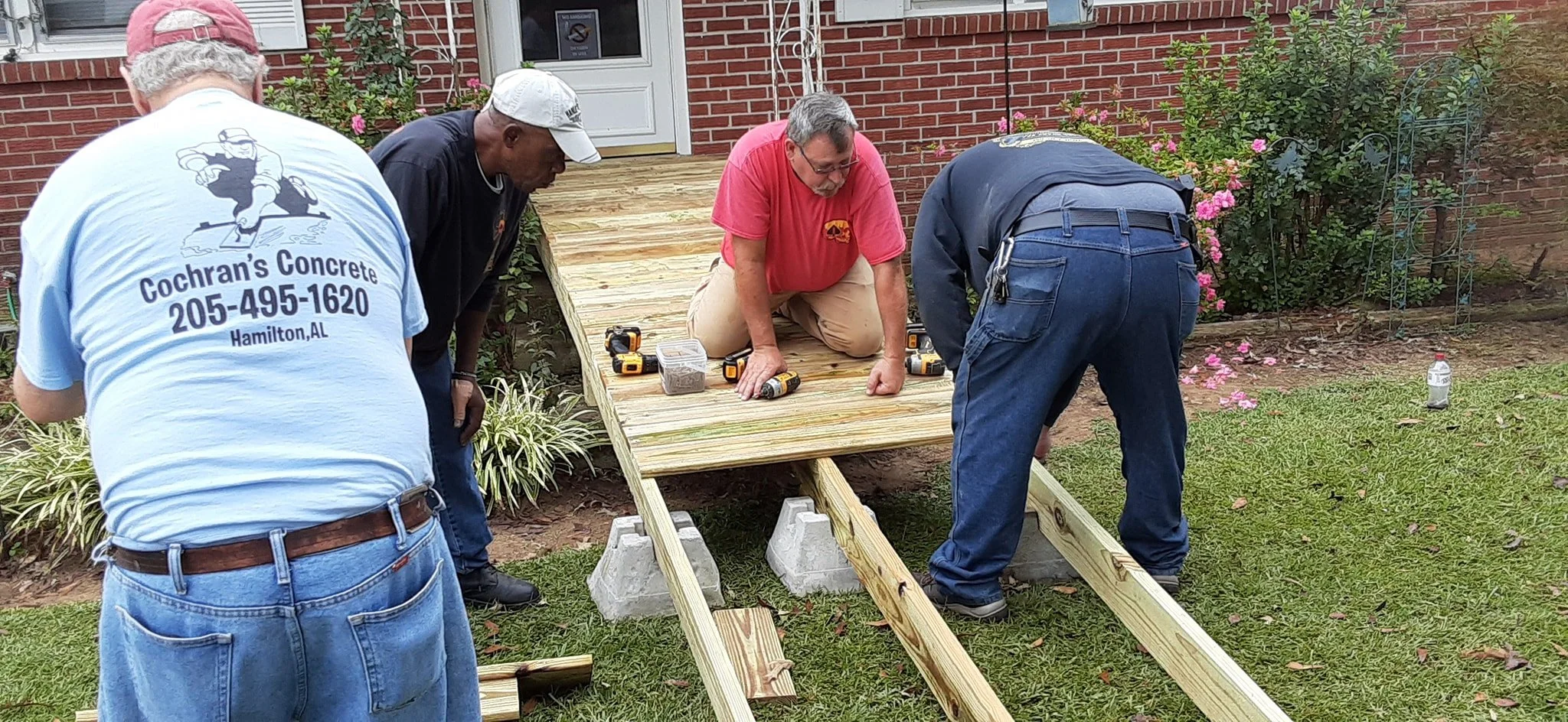 Four men working together to build a wooden ramp outside a brick house. Tools and materials are visible on the ramp, and the men are focused on the construction.