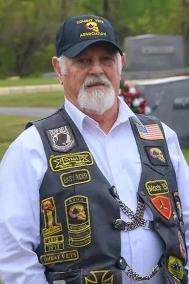 An older man wearing a black hat with gold lettering, a white shirt, and a black vest covered with various patches and badges. He is outdoors near a cemetery.