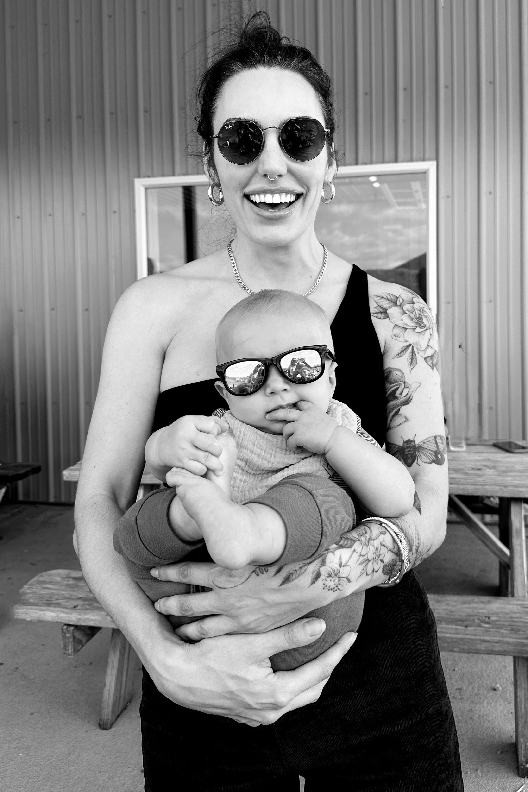 Louisiana Certified Birth Doula with tattoos and a trendy aesthetic smiles and laughs joyfully while holding her best friend's baby, both wearing sunglasses and enjoying the scenery outdoors.