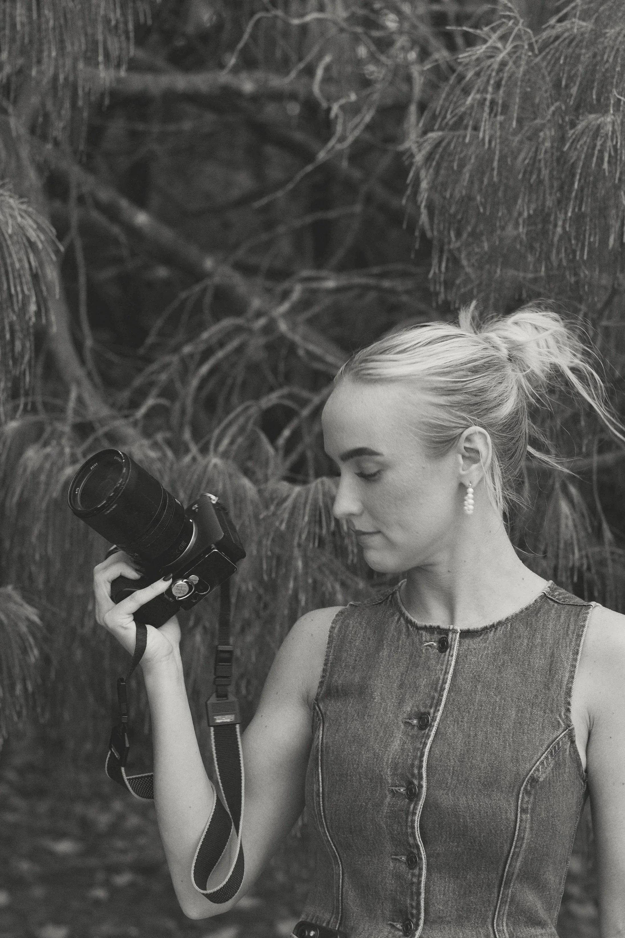 A woman with blonde hair tied in a ponytail holding a camera, standing outdoors near trees.