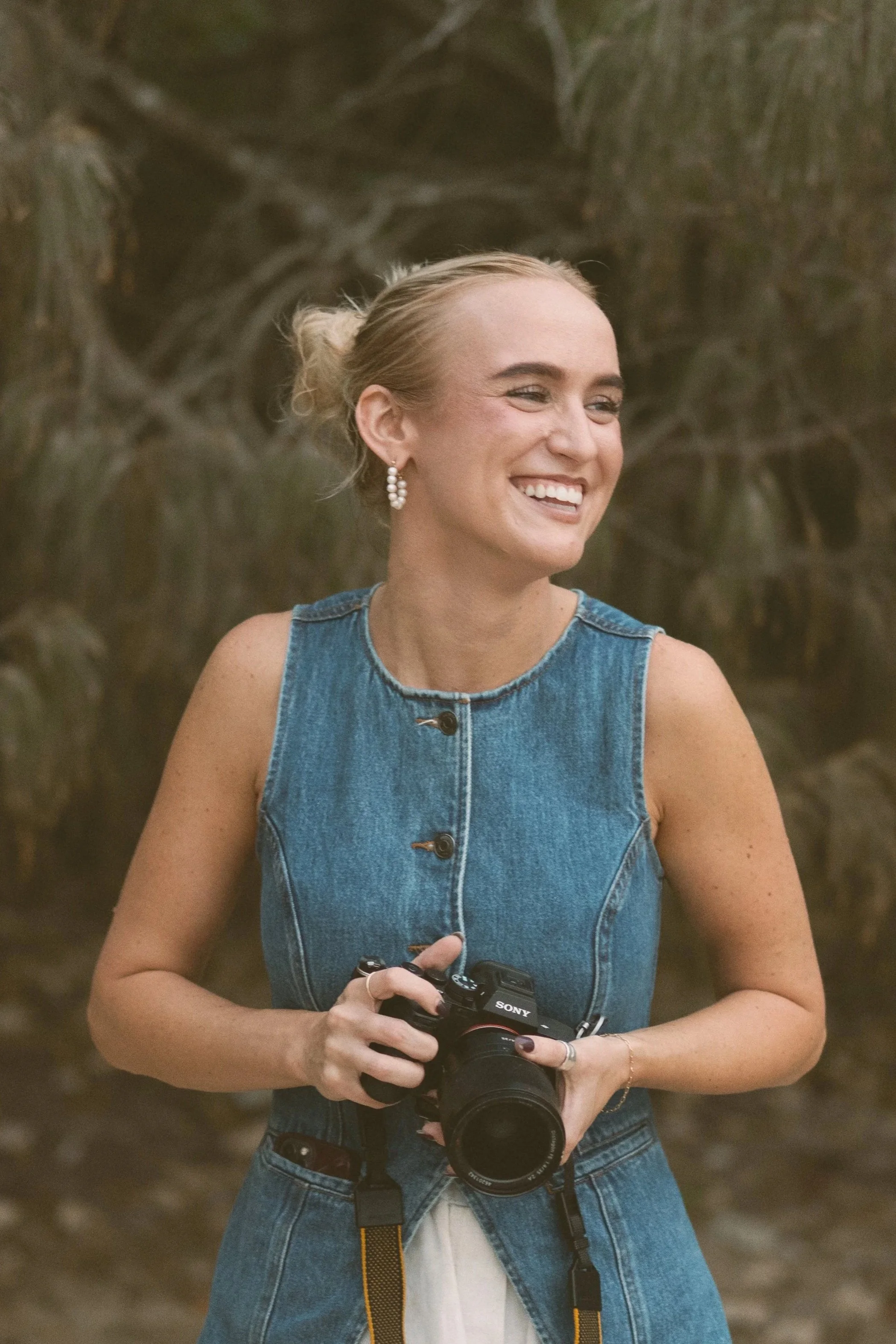 A woman smiling outdoors holds a Sony camera, wearing a sleeveless denim top and pearl earrings.