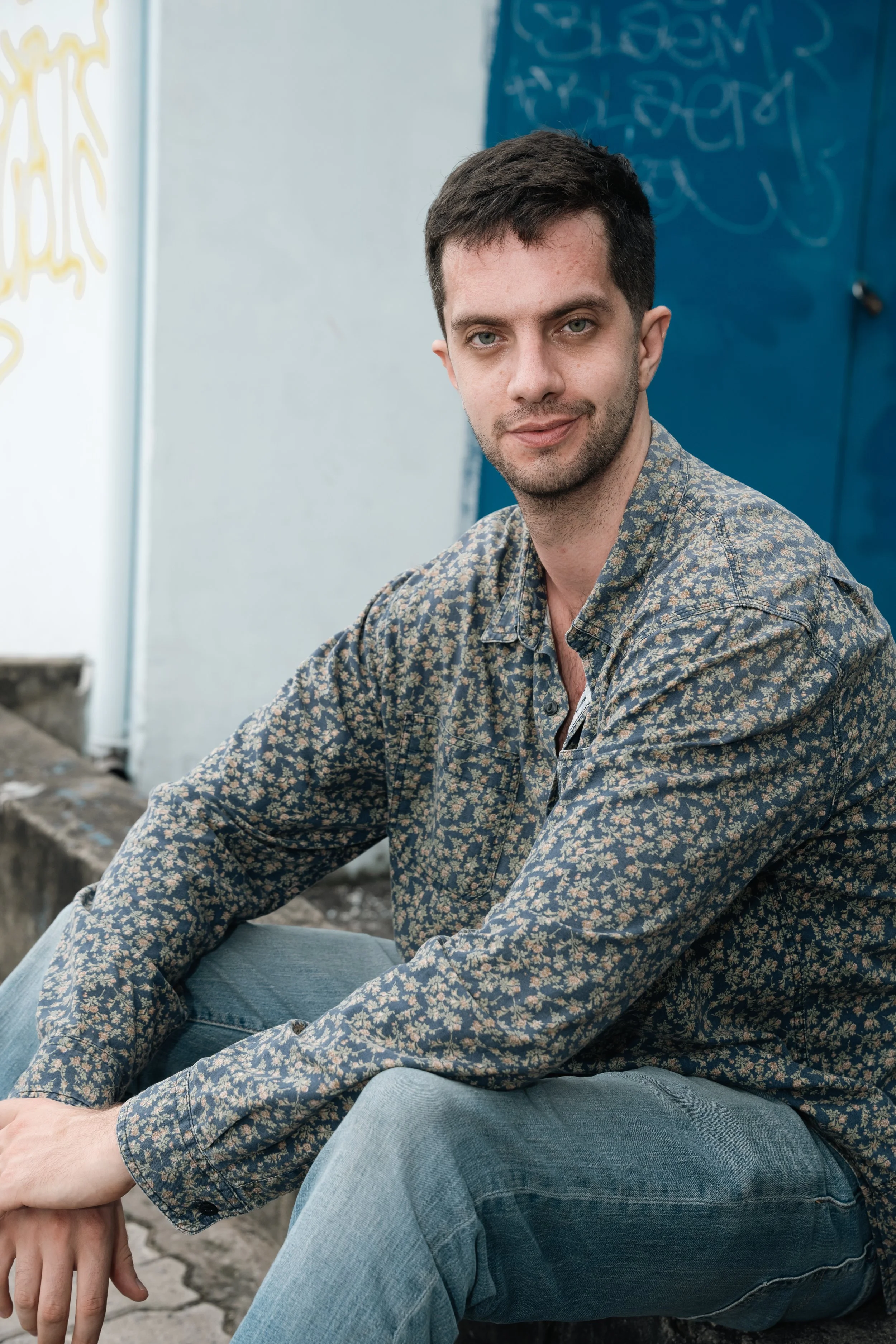 A young man with dark hair, a beard, and a light skin tone sitting outdoors on a city street curb, wearing a long-sleeve shirt with a floral pattern and blue jeans, looking at the camera with a slight smile.