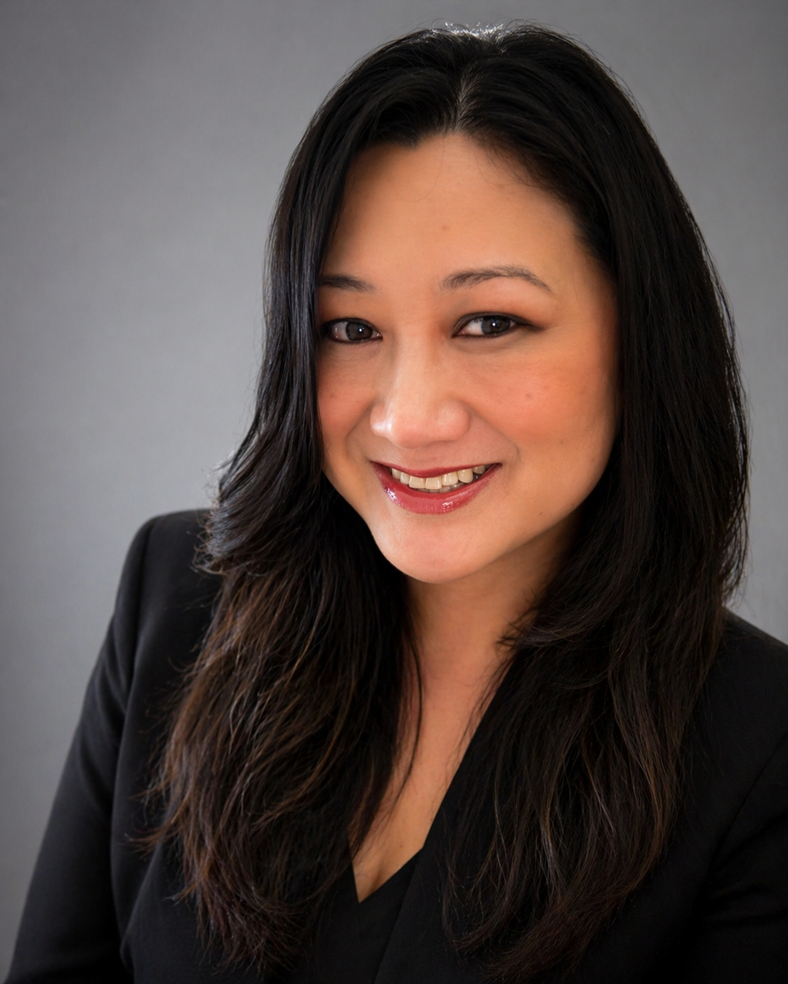 Professional woman with long dark hair smiling, wearing a black blazer, against a plain gray background.
